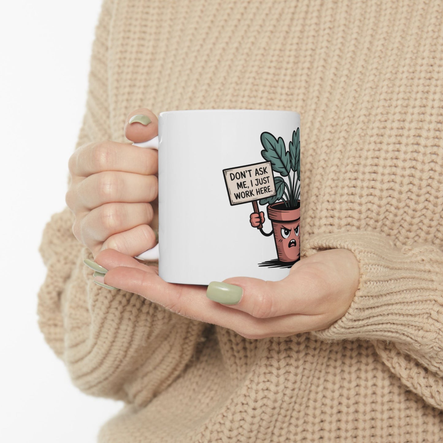 White mug with a frowning plant in a pot holding a sign that reads "DON'T ASK ME, I JUST WORK HERE."