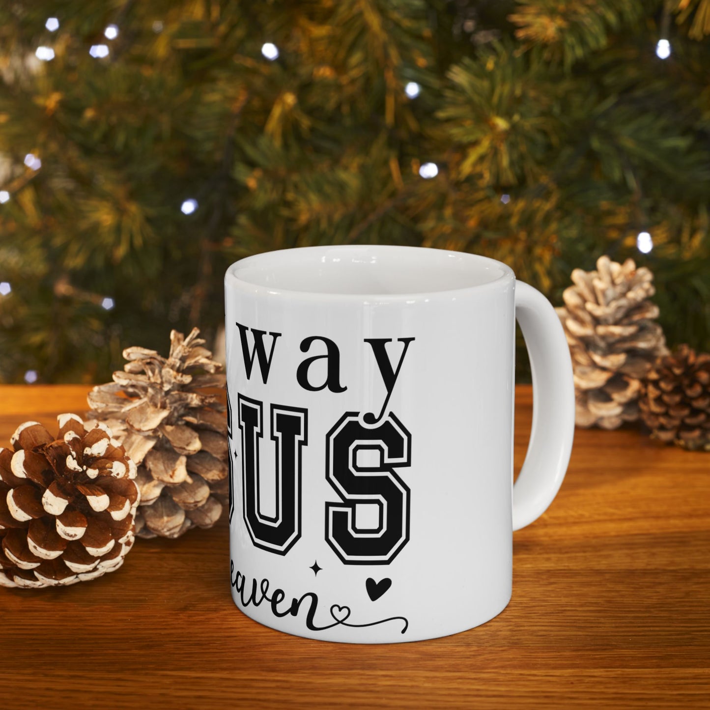 White mug with "way us heaven" text, decorated with pinecones and festive lights.