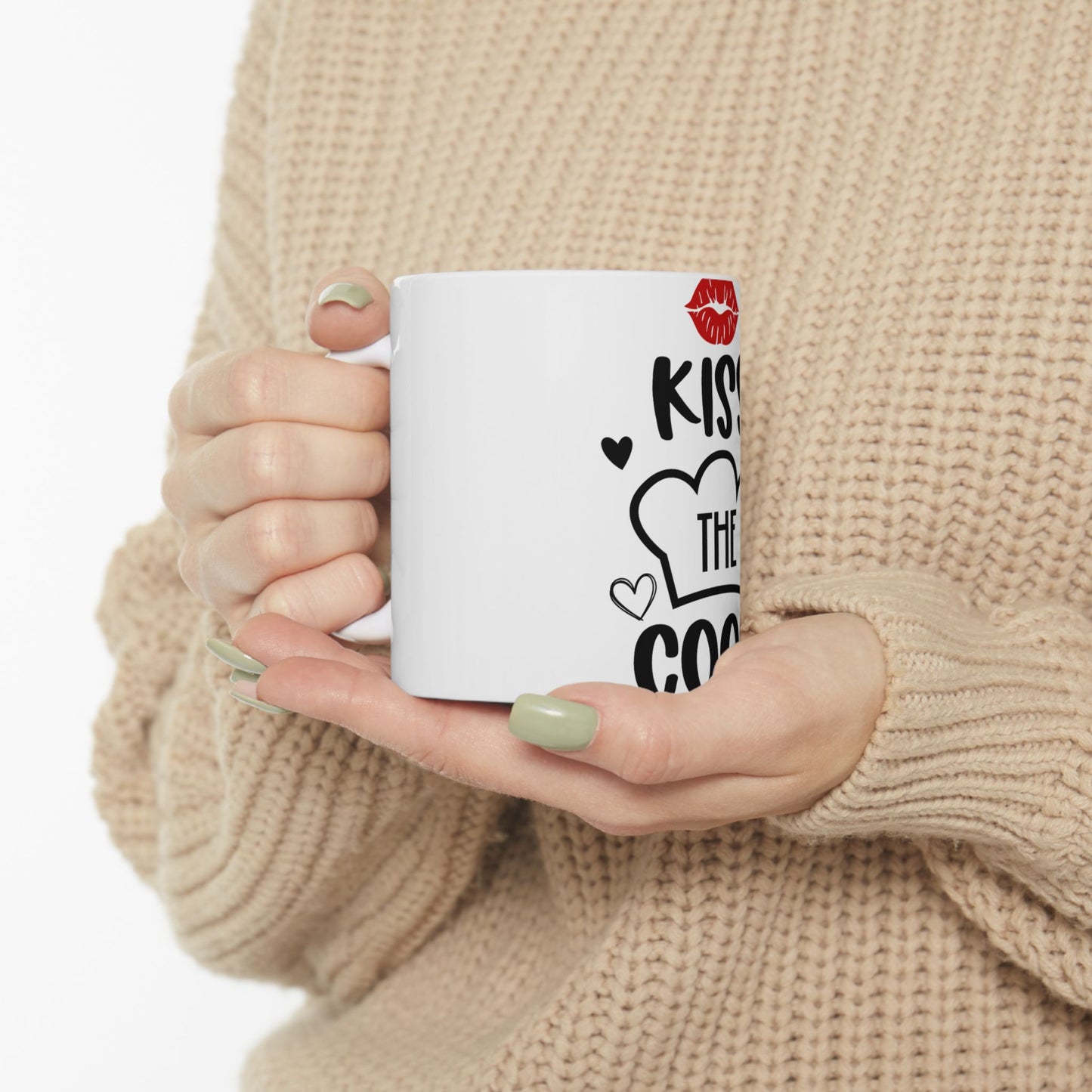 White mug with "Kiss the cook" text and heart graphics held by hands.