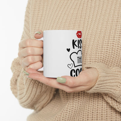 White mug with "Kiss the cook" text and heart graphics held by hands.
