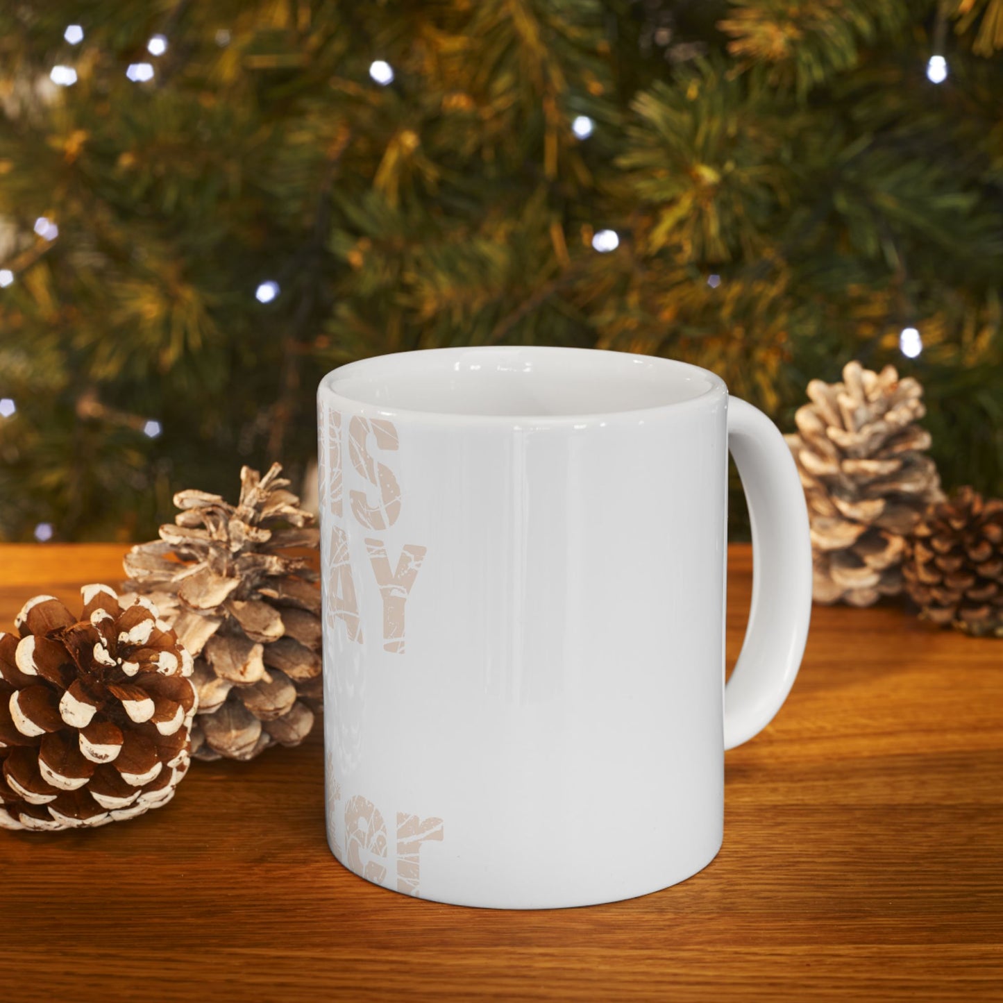 White ceramic mug with text, displayed with pinecones and a blurred Christmas tree.