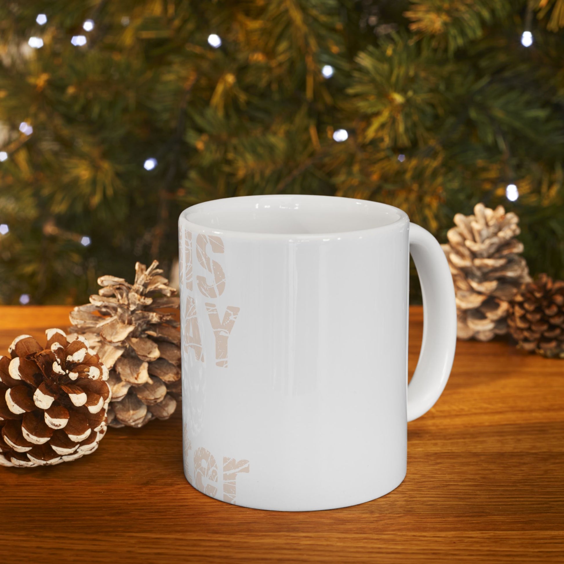 White ceramic mug with text, displayed with pinecones and a blurred Christmas tree.