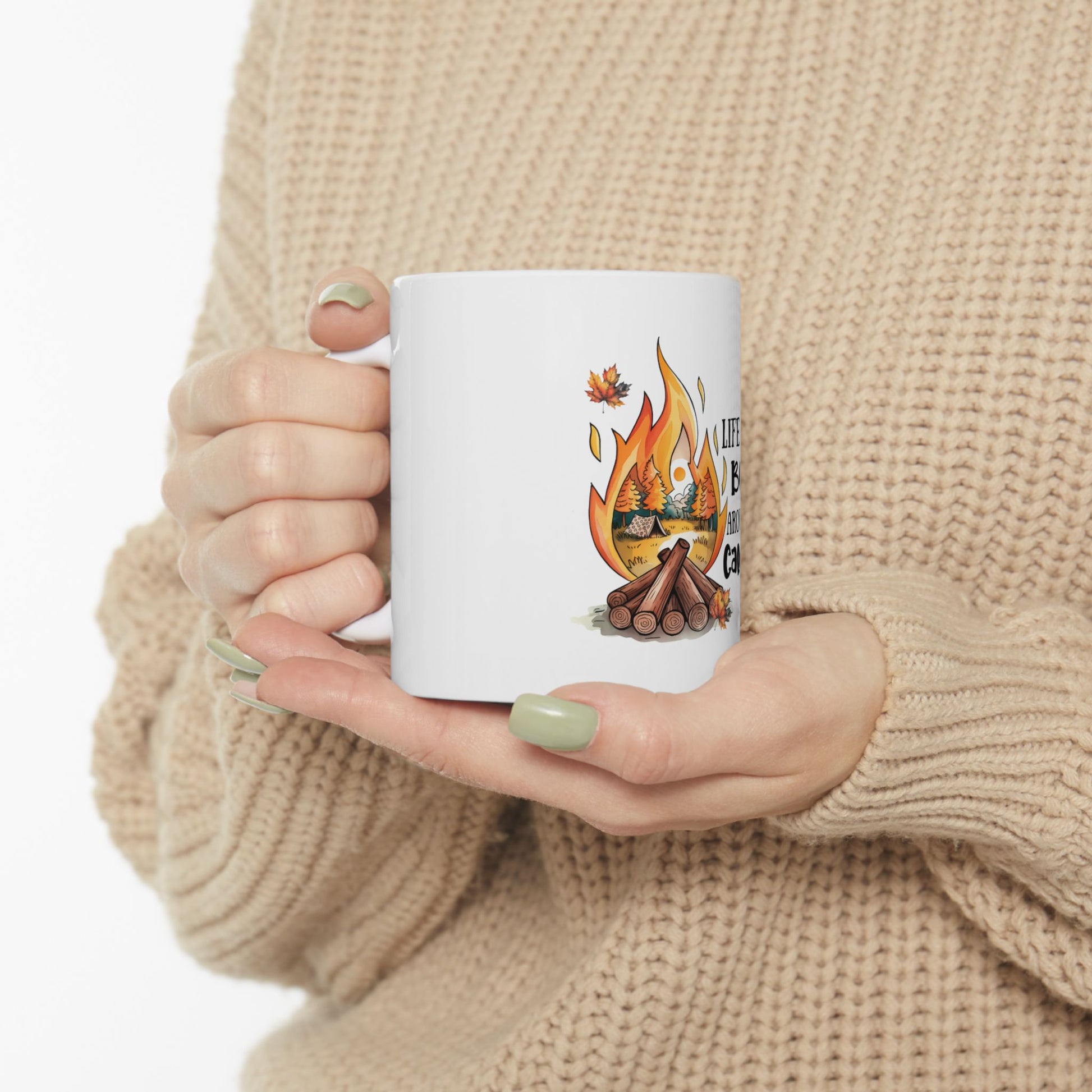 White mug with campfire illustration and text, held by person in sweater.