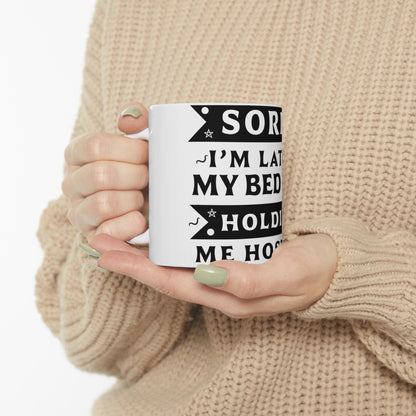 White mug with black text: "Sorry, I'm late. My bed holds me hostage."