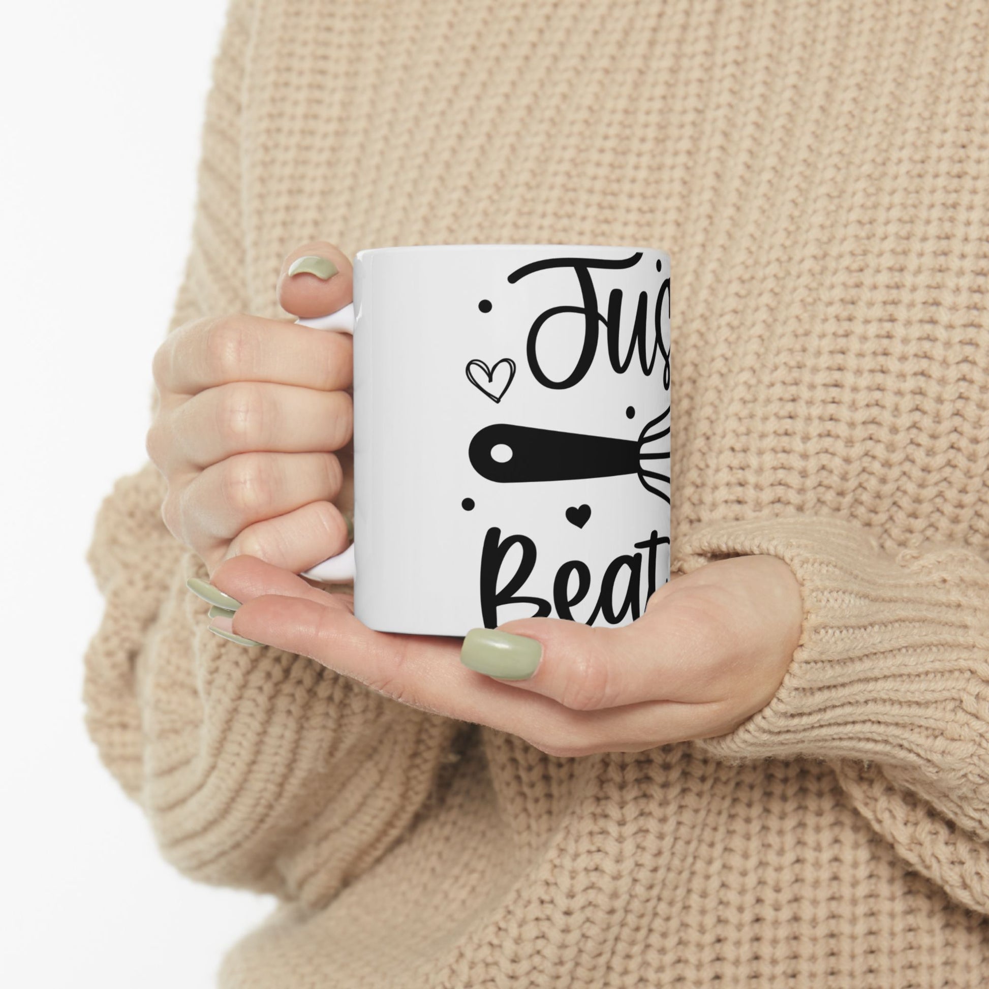 White mug with "Just Beat It" and whisk illustration held by hands.