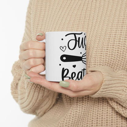 White mug with "Just Beat It" and whisk illustration held by hands.