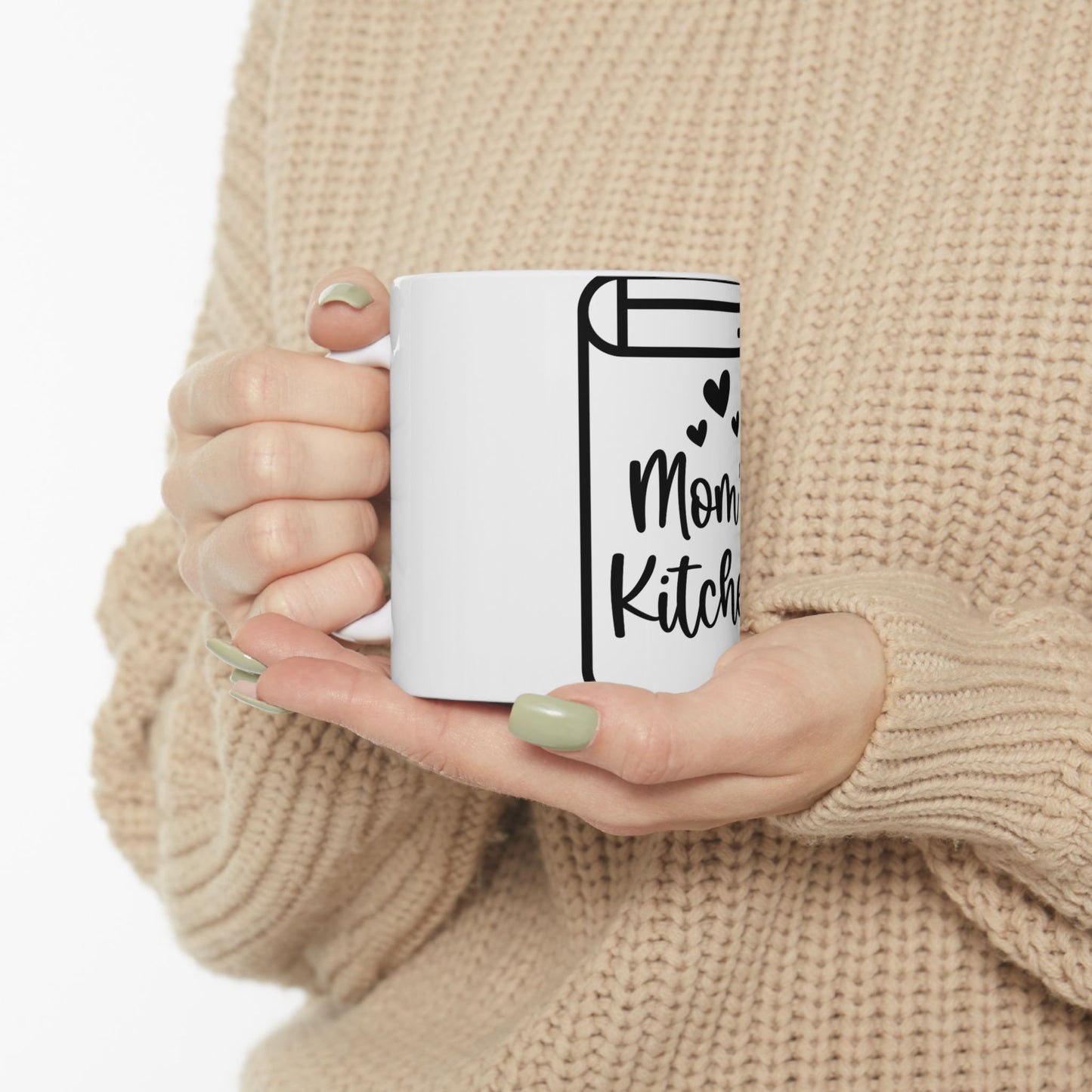 White mug with "Mom's Kitchen" text and heart accents held by hands.