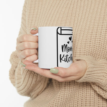 White mug with "Mom's Kitchen" text and heart accents held by hands.