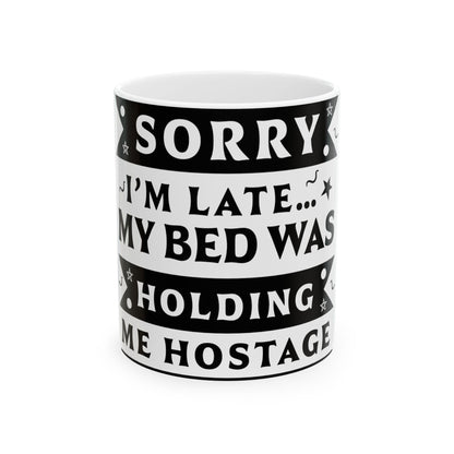 White mug with black text: "SORRY I'M LATE... MY BED WAS HOLDING ME HOSTAGE."