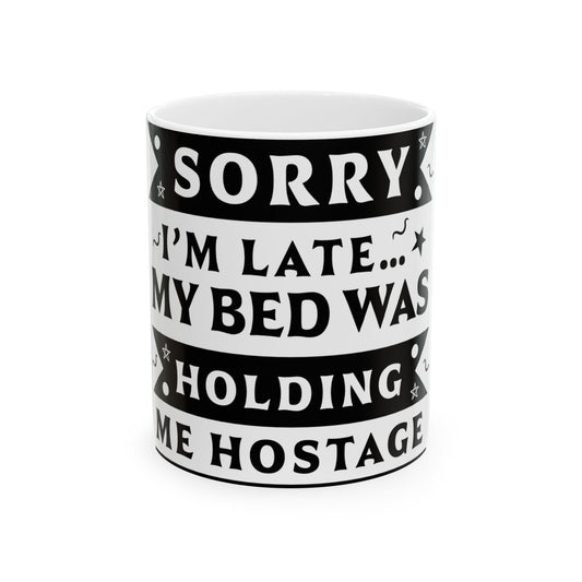 White mug with black text: "SORRY I'M LATE... MY BED WAS HOLDING ME HOSTAGE."